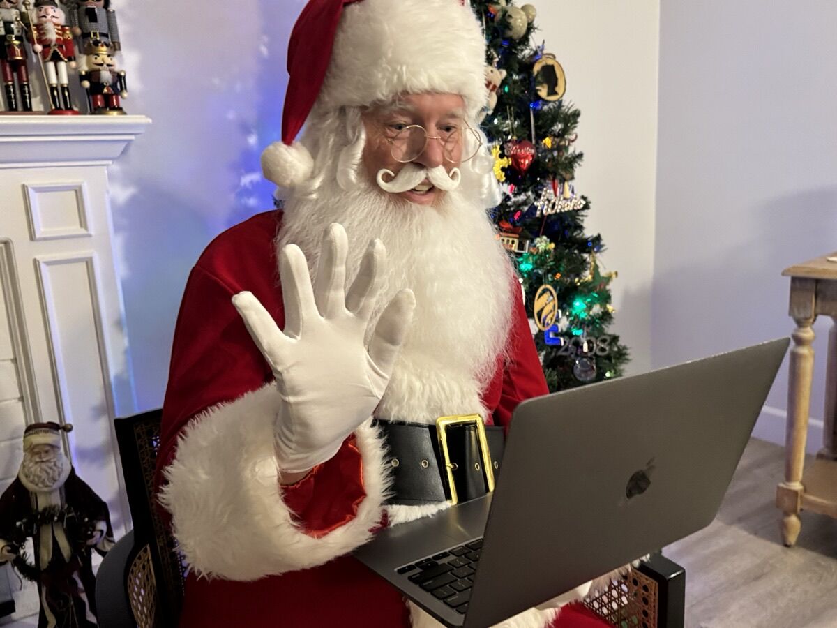 Santa in a velvet suit waving during a live video call on his laptop